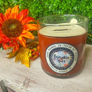 A clear glass candle with a brownish-hue and a label reading 'Bumpkin Spice'. It is displayed outdoors with fall leaves and a sunflower in the background.