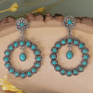 A pair of silver-toned Southwestern-style dangle earrings with turquoise stones, displayed on a rustic wood slice background with yellow flowers.