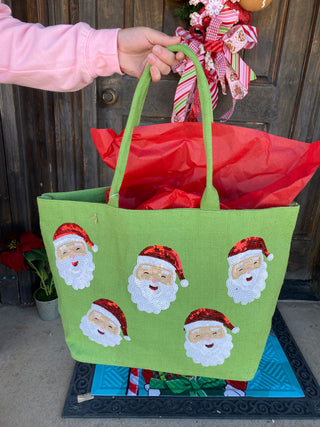 Green tote bag with Santa Claus designs held by a person in front of a door with Christmas decorations.