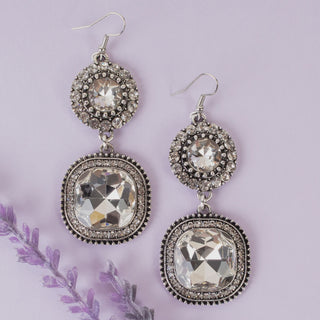 A pair of silver-toned dangle earrings with clear faceted crystals, displayed on a light lavender background with a decorative purple flower stem.