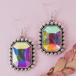 A pair of rectangular dangling earrings with faceted iridescent gemstones in silver-toned bezel settings, displayed on a soft pink background with dried pink petals at the corner.