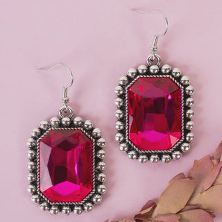 A pair of rectangular drop earrings with faceted hot pink gemstones in silver-toned bezel settings, displayed on a soft pink background with dried leaves in the corner.