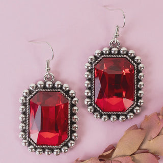 A pair of dangling earrings with large faceted red rectangular gemstones in silver-toned metal bezels, displayed on a soft pink background with dried flower petals.