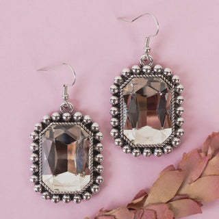 A pair of silver-toned drop earrings with large faceted clear crystal rectangular pendants, set in ornate beaded rectangular frames, displayed on a light pink background with dried pink leaves in the corner.