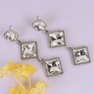 A pair of silver-tone drop earrings with clear faceted square crystals, displayed on a light lavender background with yellow flowers in the corner.