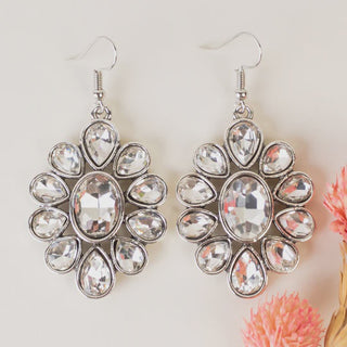 A pair of silver-tone flower-shaped drop earrings featuring a large oval crystal center surrounded by teardrop-shaped clear crystals, displayed on a white surface with pink accents.