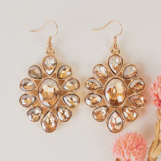 A pair of gold-tone flower-shaped drop earrings featuring a large oval champagne crystal center surrounded by teardrop-shaped crystals, displayed on a white surface with pink accents.