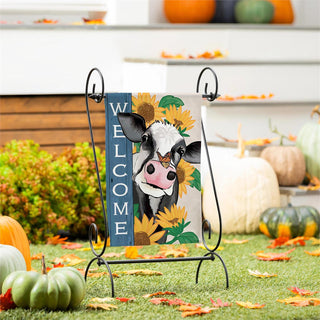 A decorative garden flag with a cow face, sunflowers, and a blue vertical banner reading “WELCOME,” displayed on a black metal stand in an outdoor fall yard with pumpkins and leaves.
