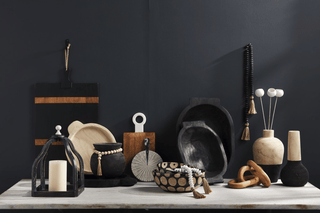 A styled collection of modern home decor and tableware displayed on a light marble surface against a dark charcoal wall. The collection includes matte black ceramic vessels, natural wood cutting boards, a black lantern with a white pillar candle, patterned bowls, and minimalist vases.