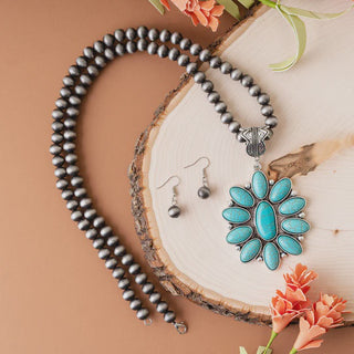 A jewelry set featuring a turquoise blossom pendant on a strand of burnished silver beads, paired with matching silver bead drop earrings, displayed on a wooden slice.