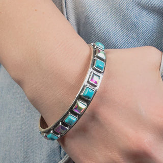 A silver cuff bracelet featuring alternating square turquoise stones and iridescent crystals, worn on a wrist.