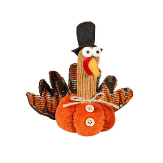 A handmade Thanksgiving turkey decoration made from a knitted sock turkey body wearing a black top hat, with orange and brown patterned feathers, a red and yellow beak, and an orange fabric pumpkin base with two buttons.