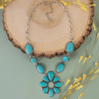 A western-style silver chain necklace featuring oval turquoise stones leading to a large turquoise squash blossom pendant, displayed on a rustic wood slice.