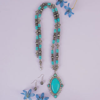 A turquoise and silver beaded necklace with a large oval turquoise pendant, displayed on a light lavender background with matching silver and turquoise earrings and small blue flower props.