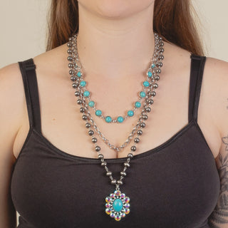 A close-up of a woman wearing a multi-strand necklace. The necklace features silver-toned beaded strands, a shorter strand with turquoise blue oval stones, and a long pendant necklace with a turquoise blue oval center surrounded by iridescent multicolor stones.