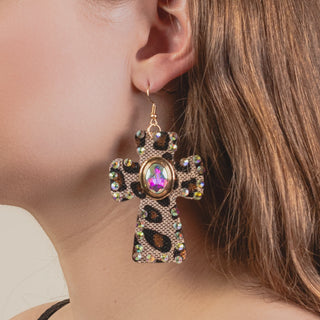 A close-up of a person wearing a large, leopard-print cross earring embellished with iridescent crystals and a central pink-toned gemstone.