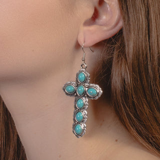 A close-up of a person wearing a large, southwestern-style silver cross earring set with six oval turquoise stones and detailed engraved borders.