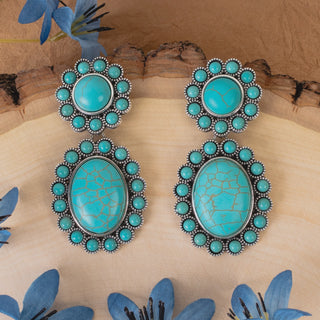 A pair of silver-toned drop earrings with oval turquoise cabochons and circular turquoise accents, displayed on a light wood surface with blue flowers and bark in the background.