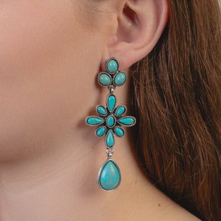 A close-up of a woman's ear wearing a dangling turquoise earring with silver-toned metal detailing and a teardrop-shaped drop stone.