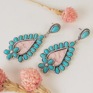 A pair of teardrop-shaped dangle earrings featuring light pink textured centers, bordered by turquoise stones and a small turquoise flower accent, shown on a white surface with pink dried flowers.