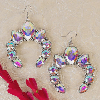 A pair of silver-tone crescent-shaped earrings featuring a variety of teardrop and round iridescent aurora borealis crystals, shown on a textured gold background with red petals.
