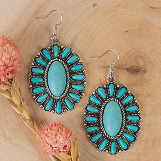 A pair of silver-tone oval sunburst earrings featuring a large center turquoise stone surrounded by a halo of smaller turquoise cabochons, displayed on a wooden slice with pink dried flowers.