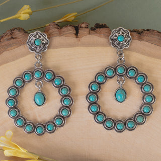 A pair of silver-toned Southwestern-style dangle earrings with turquoise stones, displayed on a rustic wood slice background with yellow flowers.
