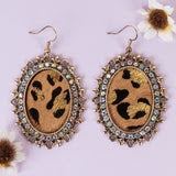 A pair of oval leopard-print drop earrings featuring a scalloped antique gold-tone border embellished with small iridescent crystals, shown on a light purple background with white flowers.