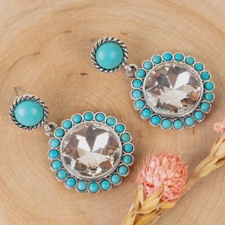 A pair of silver-tone dangle earrings with round turquoise blue beads and large clear faceted center stones, displayed on a light wood surface with dried pink flowers.