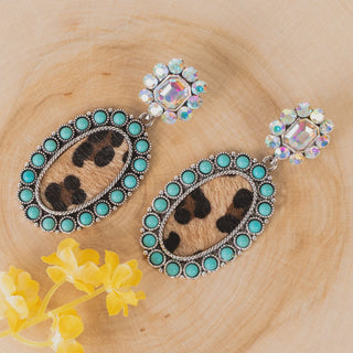 A pair of bohemian dangle earrings with oval turquoise and leopard print hair-on-hide centers, set in silver-tone filigree with iridescent crystals at the studs, displayed on a light wood background with yellow flowers.