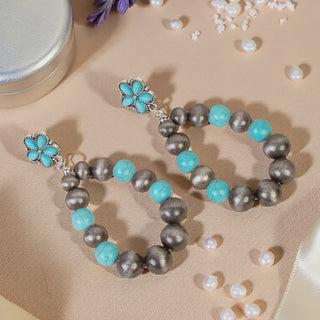 A pair of teardrop-shaped hoop earrings featuring burnished silver beads and turquoise accents, hanging from a small turquoise flower stud, displayed on a tan surface with scattered pearls.
