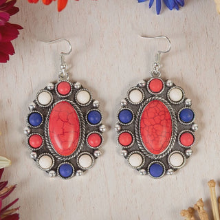 A pair of oval silver-tone drop earrings featuring a large red faux-stone center, surrounded by a halo of smaller red, white, and blue cabochons in a patriotic theme, displayed on a light wood surface