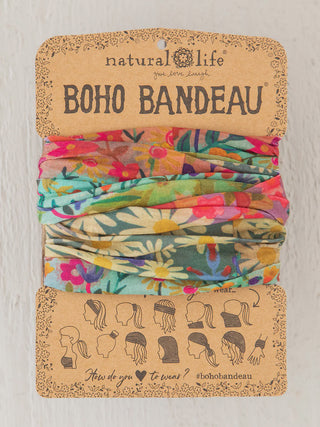 This Natural Life "Boho Bandeau" features a vibrant, garden-inspired print with yellow daisies and colorful wildflowers against teal and tan backgrounds. The fabric is displayed on its original cardboard packaging, which highlights the product's versatility through a series of illustrated "how-to-wear" icons.