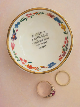 Decorative ring dish with floral design and text, placed on a pink surface 