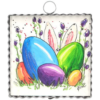 A small, square, metal-framed piece of art featuring a watercolor drawing of several brightly colored Easter eggs (blue, green, purple, orange) nestled in grass with purple flowers, and two white bunny ears with pink insides visible behind the eggs
