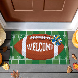 A football-themed doormat with a brown football graphic, white laces, the word 'WELCOME' in white, and a blue and yellow pom-pom, placed in front of a brown door on a gray deck with pumpkins and autumn leaves around it.