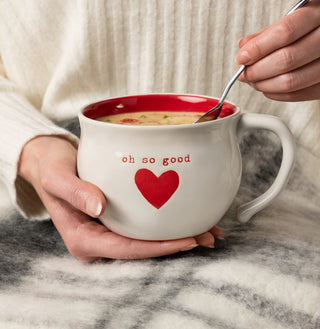A person holding a white ceramic soup bowl with a red heart and the text 'Oh So Good' on it.