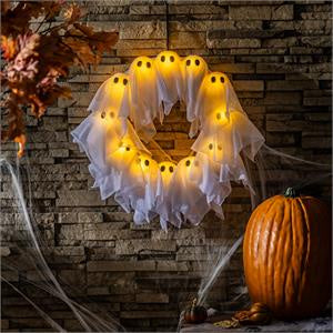 A Halloween wreath decoration made of white ghost fabric with glowing yellow faces, hanging on a rustic stone wall beside a pumpkin and cobwebs.