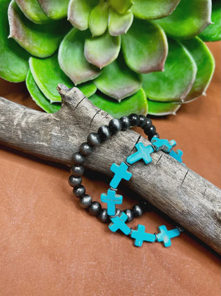 Two beaded bracelets displayed on a weathered branch: one with dark gray beads and the other with turquoise cross charms, set against a brown surface with a green succulent in the background.