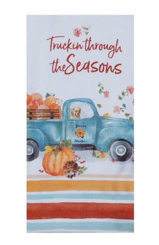 A rectangular kitchen towel featuring a watercolor illustration of a blue pickup truck carrying pumpkins, with the phrase 'Truckin through the Seasons' in red script. The towel has a white base with a decorative autumn-themed design and striped accents at the bottom.