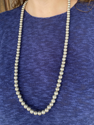 A long strand of light gray faux pearls worn as a necklace over a textured blue knit top.