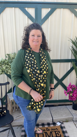 Woman wearing a green sweater and gold shamrock-patterned on green background scarf standing on a porch.