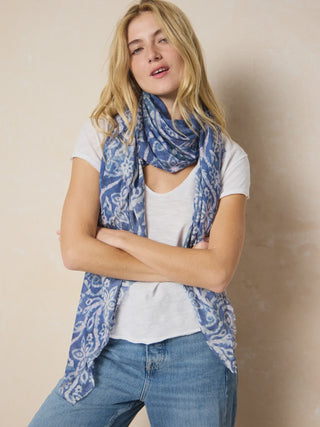 Woman wearing a blue patterned scarf, white shirt, and jeans against a beige background
