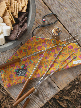 Barbecue tools with wooden handles on a floral-patterned mat next to a bowl of graham crackers, chocolate, and marshmallows.