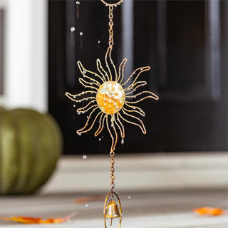 A decorative autumn sun wind spinner with a gold wireframe sun, a glittery amber center, and a small hanging bell, shown hanging in front of a window.