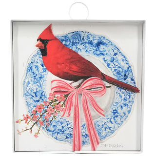A red cardinal sitting on a branch with cherry blossoms, a pink bow tied around the branch, on front of a blue and white chinoiserie print