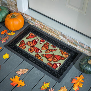 A rectangular doormat with a black ornate border and a central design of orange and red monarch butterflies on a muted green background, shown on a gray deck in front of a door with pumpkins and autumn leaves around it.