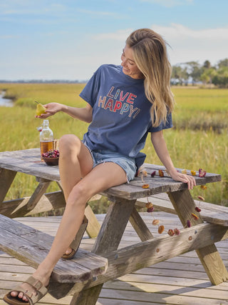 Woman sitting at a picnic table with a 'Live Happy' shirt, surrounded by nature.
